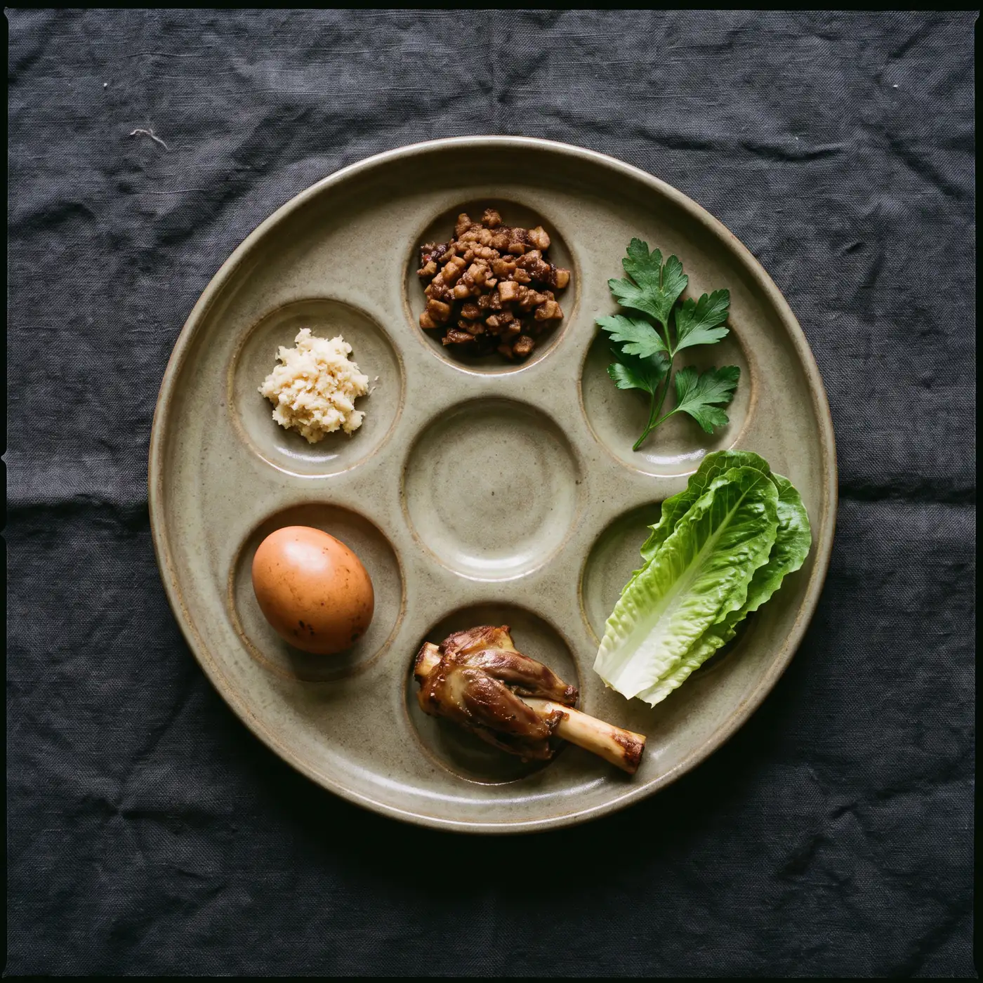 A seder plate seen from directly above, its wells filled with shank bone, roasted egg, parsley, charoset, horseradish and romaine